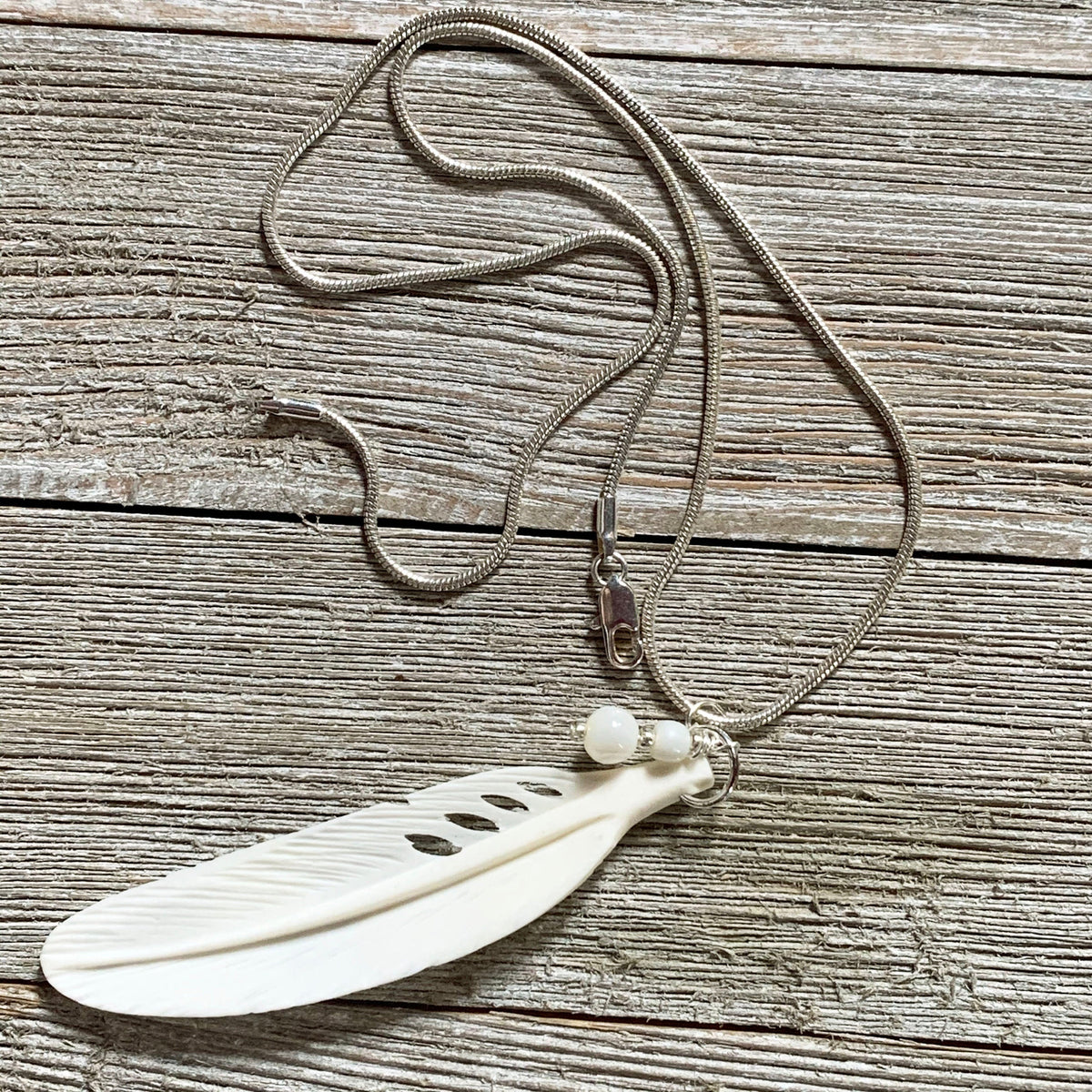 The White Feather Necklace features an angel sign carved bone and mother of pearl pendant, displayed on a rustic wooden surface—ideal for those seeking meaningful spiritual jewelry.