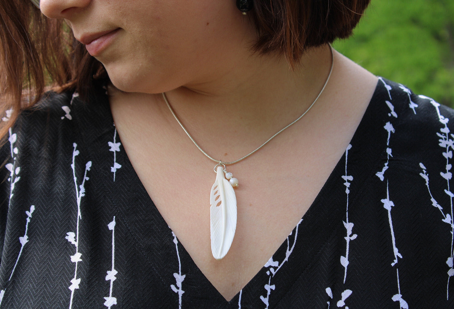 A woman wearing a black top with a white branch pattern and the White Feather Necklace: Mother of Pearl Pendant, 18-inch Chain, is photographed outdoors with greenery in the background.