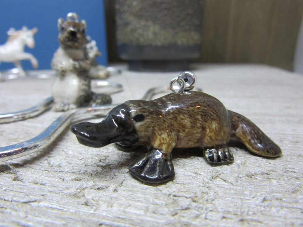 The Exotic Porcelain Platypus Figurine Bookmark for Book Reader Gift, featuring a handy metal loop, rests on wood as an ideal whimsical page marker. A blurred ceramic animal and silver object enhance the scene&