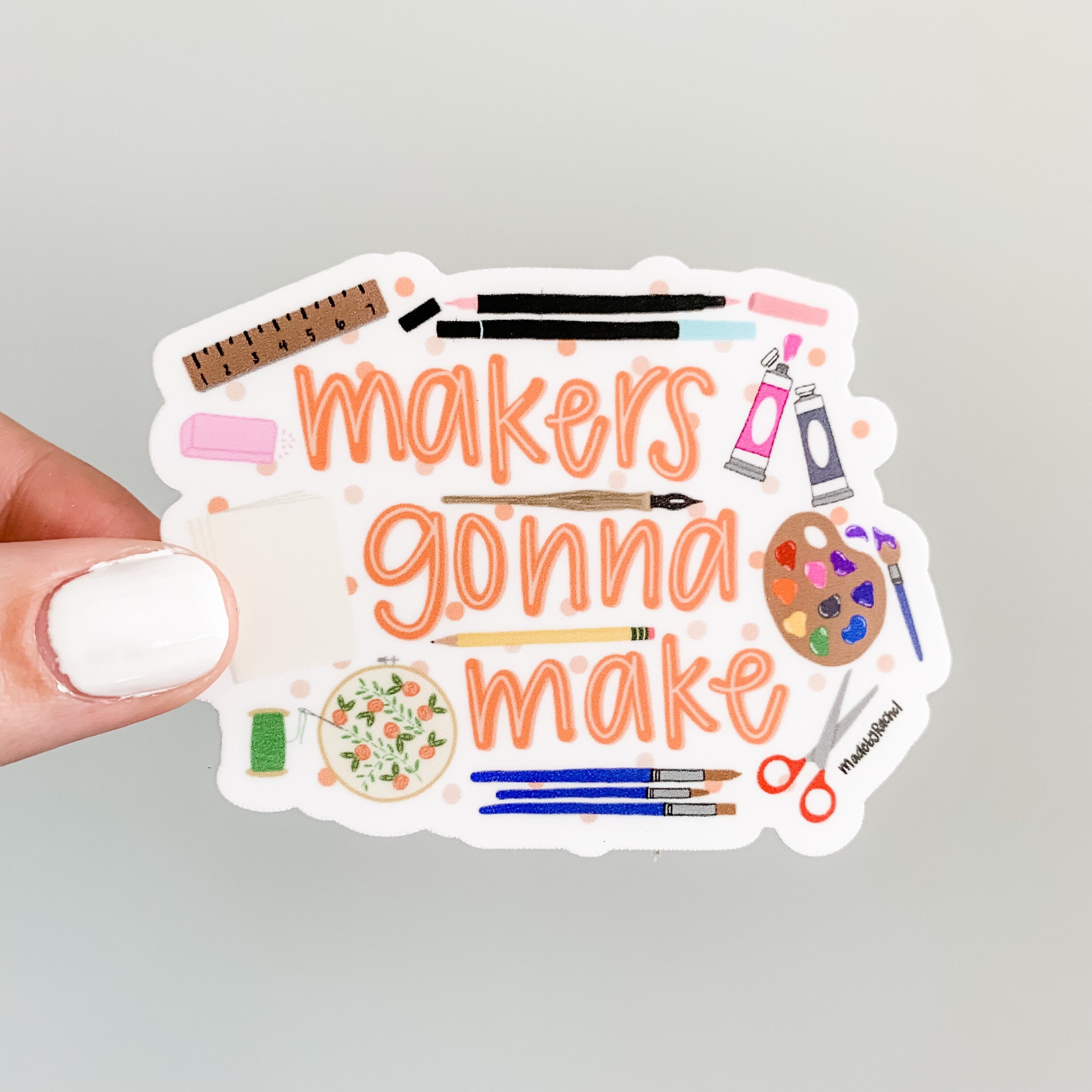 the phrase makers gonna make handwritten in orange, surrounded by illustrations of a ruler, markers, paint, a paint palette and brush, scissors, writing utensils, an embroidery hoop, paper, and eraser, on a white background with faint polka dots