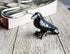 A hand-painted raven figurine bookmark rests on a wooden surface near the edge of an open book, alongside a unique metal book lover gift shaped like a hook.