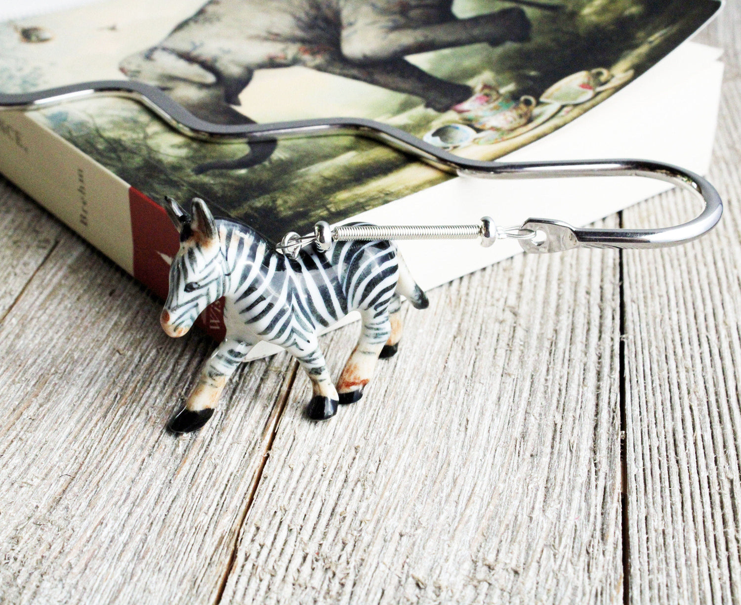 A zebra-shaped keychain hook sits on wood beside a book with an animal cover, making it a fun reading gift or a perfect match for the Porcelain Zebra Bookmark – Animal Lover Gift for Readers, Book Fans.
