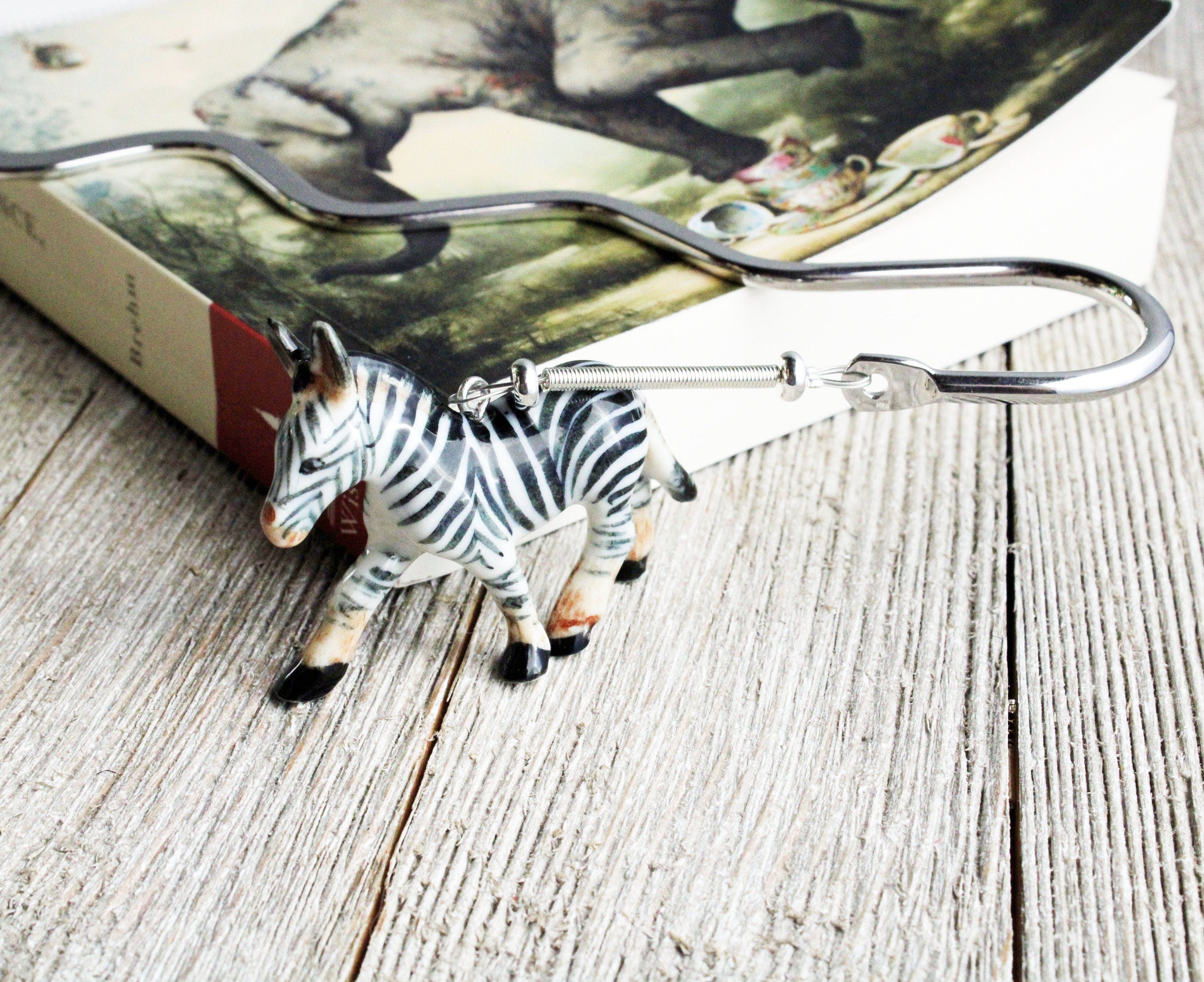 A zebra-shaped keychain hook sits on wood beside a book with an animal cover, making it a fun reading gift or a perfect match for the Porcelain Zebra Bookmark – Animal Lover Gift for Readers, Book Fans.