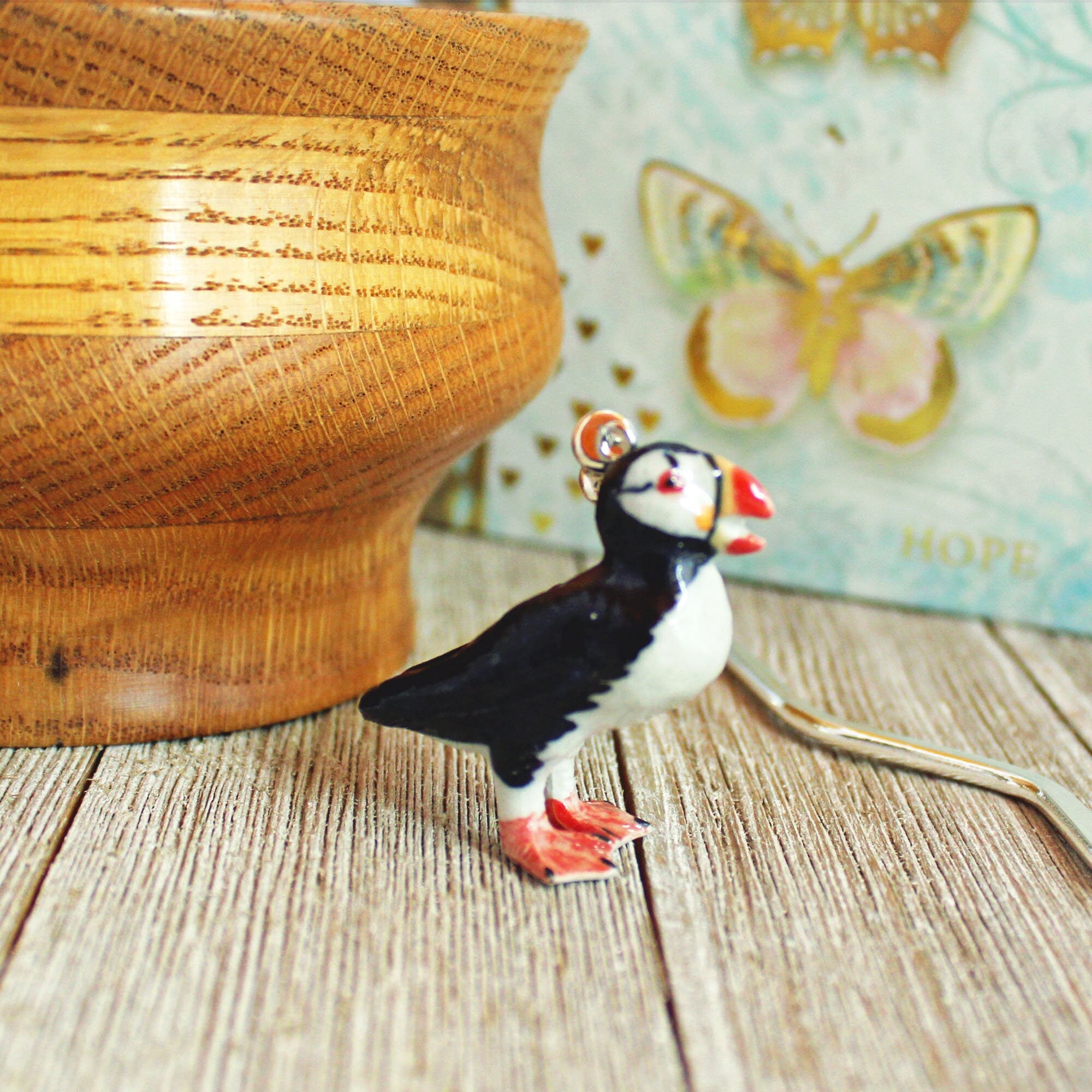 A Puffin Porcelain Bookmark – Unique Ocean Lover Gift, Handmade Figurine Bookmark, rests on a wooden surface near a decorative bowl and butterfly art, its puffin figurine featuring a red beak and feet.
