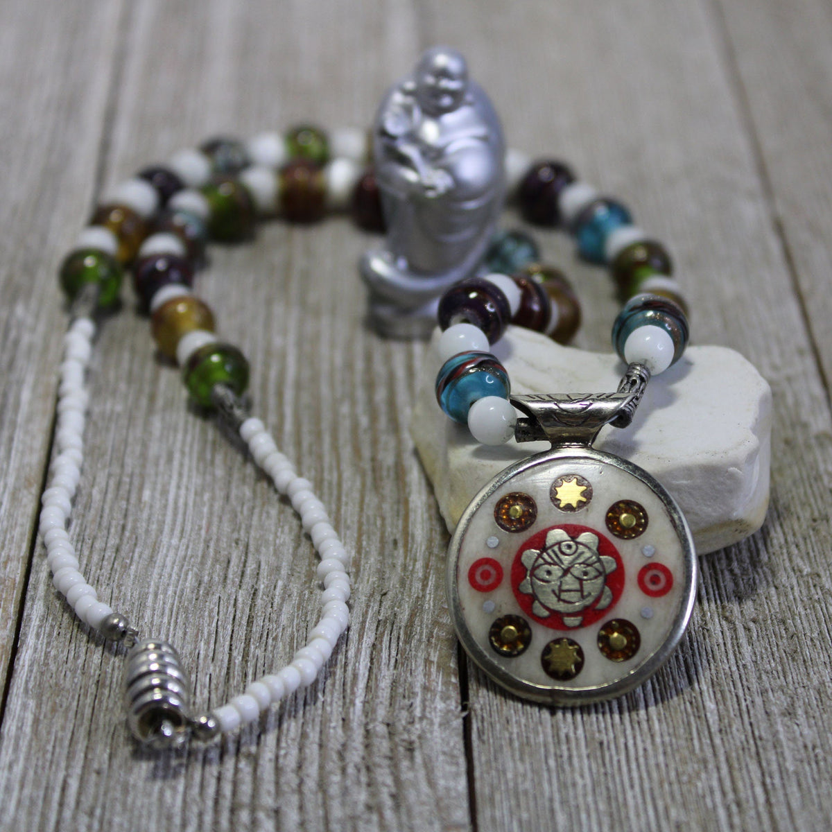 The Reversible Tibetan Pendant Necklace—crafted from vintage sterling silver with glass beads and a circular floral star design—rests on a white heart stone with a silver Buddha figurine, perfect for lovers of unique Zen jewelry.