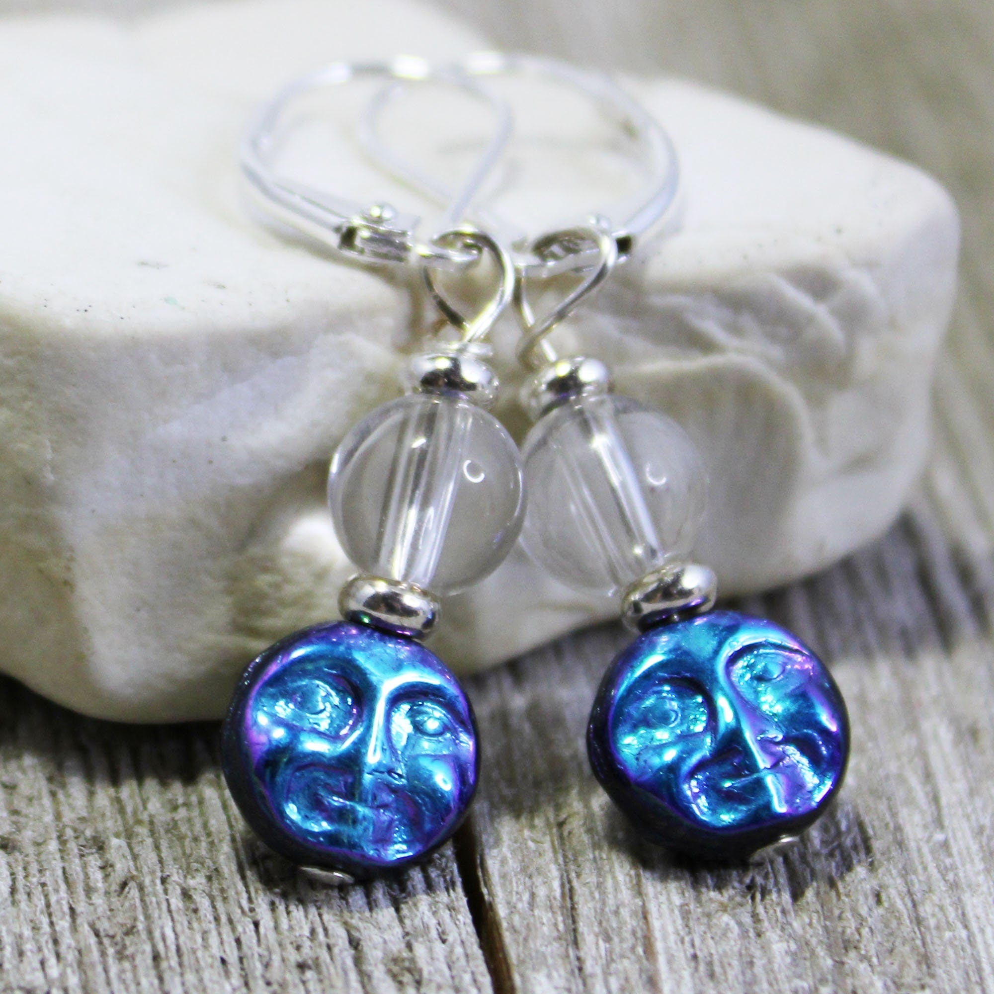 The Last Pair Full Moon Quartz Crystal Earrings feature clear round beads and blue moon face charms on sterling silver lever backs, displayed on stone with a rustic wood background—ideal for quartz crystal earring fans.