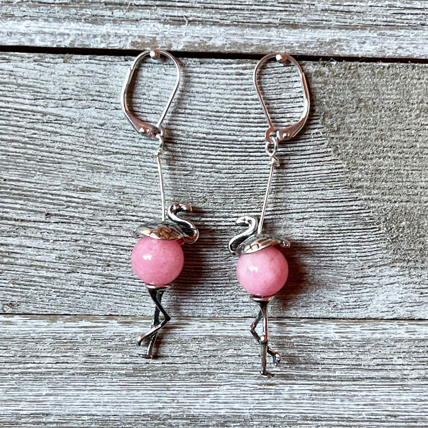 The Pink Flamingo Sterling Silver Earrings – Handmade Tropical Jewelry feature round pink beads as bodies with detailed metalwork for the head, legs, and wings, beautifully displayed on weathered wood.
