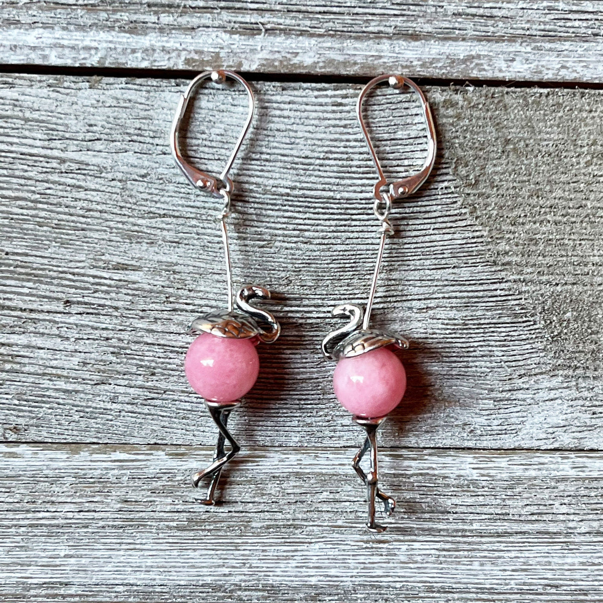 The Pink Flamingo Sterling Silver Earrings – Handmade Tropical Jewelry feature round pink beads as bodies with detailed metalwork for the head, legs, and wings, beautifully displayed on weathered wood.