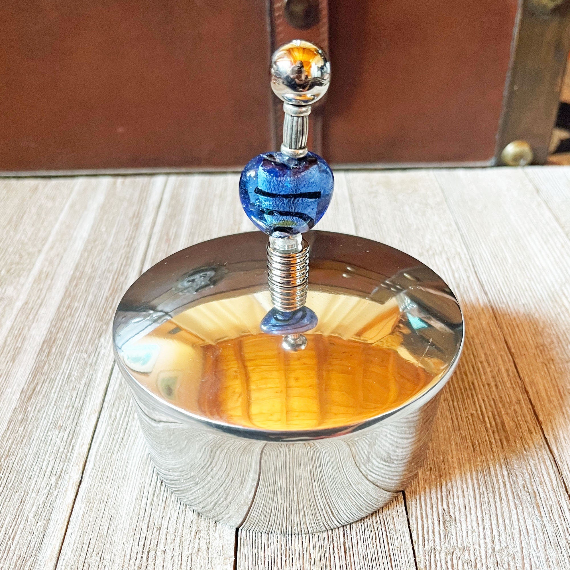 The Stainless Steel Trinket Box: Blue Foil Heart Glass Bead, Romantic Desk Decor features a shiny, round stainless steel design with a reflective surface and a handle topped with a blue foil heart glass bead—ideal for keepsake storage.