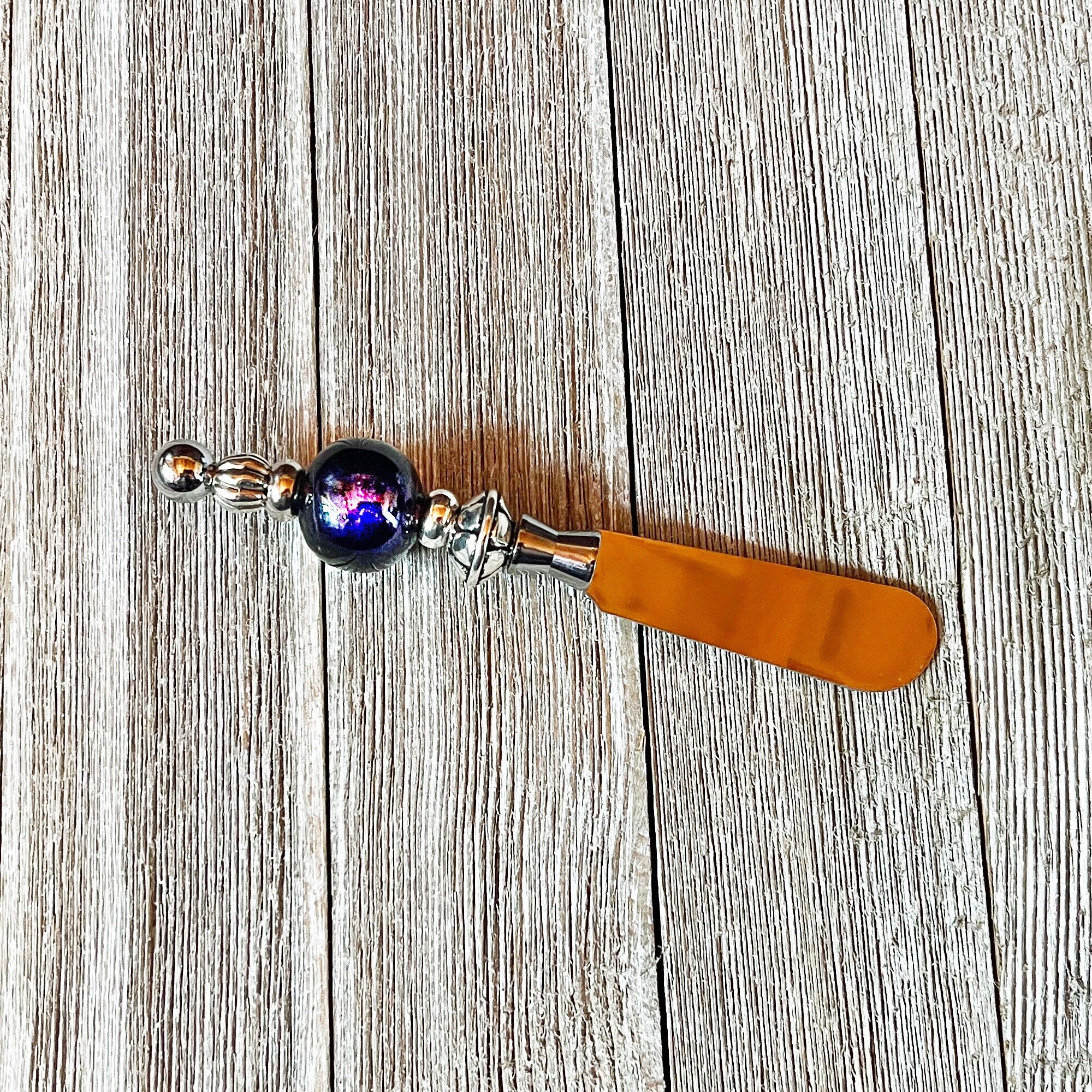 The Beaded Handle Cheese Knife, a silvertone kitchen accessory with a shiny orange blade and a round, dark blue and purple marbled accent, rests on a weathered wooden surface.