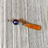 The Beaded Handle Cheese Knife, a silvertone kitchen accessory with a shiny orange blade and a round, dark blue and purple marbled accent, rests on a weathered wooden surface.