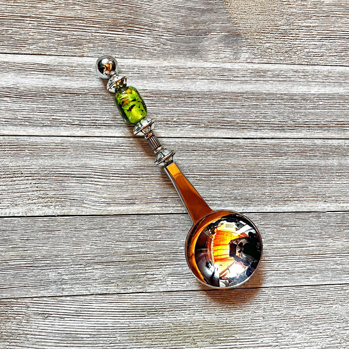 The Art Glass Beaded Handle Coffee Spoon, a stainless steel 1 Tbsp measure with a green art glass bead on its handle, rests on a textured wooden surface. The spoon’s bowl reflects colorful indoor lights and surroundings.