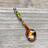 The Art Glass Beaded Handle Coffee Spoon, a stainless steel 1 Tbsp measure with a green art glass bead on its handle, rests on a textured wooden surface. The spoon’s bowl reflects colorful indoor lights and surroundings.