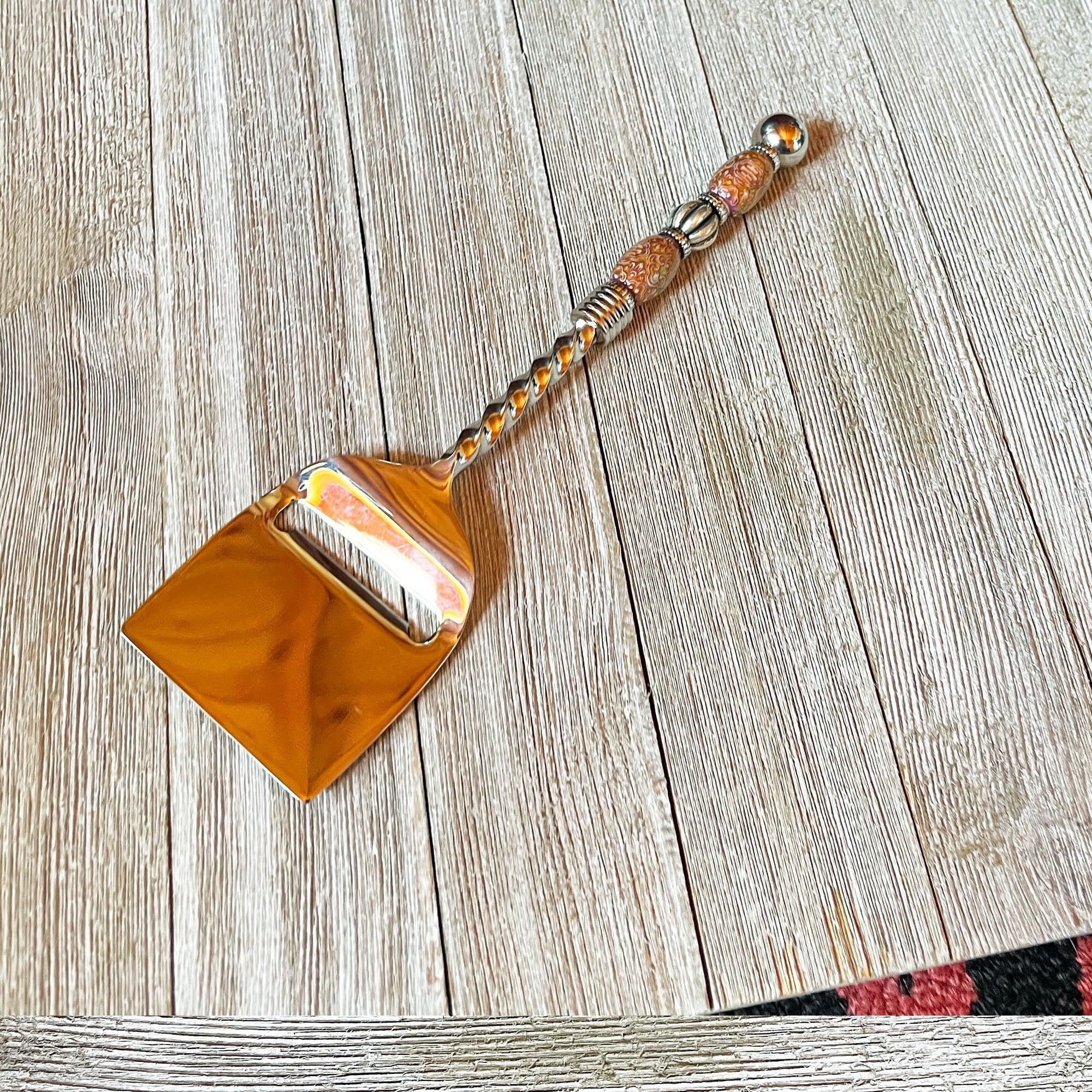 The Color Changing Beaded Cheese Slicer - Stainless Steel Party Tableware, featuring a decorative handle, sits on a light wooden surface that showcases its unique beaded design and the wood&