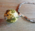 A close-up of the Ocean Jasper Atlantis Stone Orb Pendant Necklace, featuring green and yellow patterns in a floral design and metallic cylindrical beads, showcases its artisan-made minimalist style on a wooden surface.