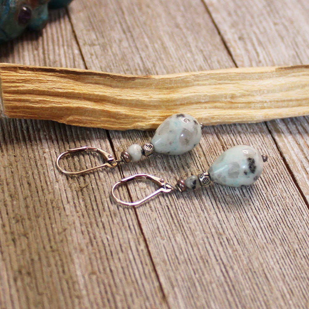 Sesame Jasper Mint Earrings with Black Tourmaline in an artisan design and sterling silver hooks, featuring sacred geometry, are displayed on a wooden surface beside a palo santo stick.