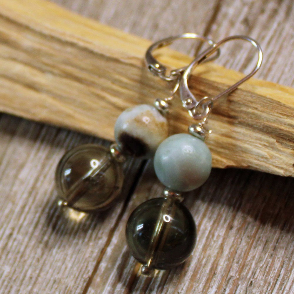 A pair of Larimar Smokey Quartz Earrings—Sterling Silver Sacred Geometry Jewelry—with round greenish-grey and light blue beads, minimalist design, and metal hooks, displayed on a wooden surface.