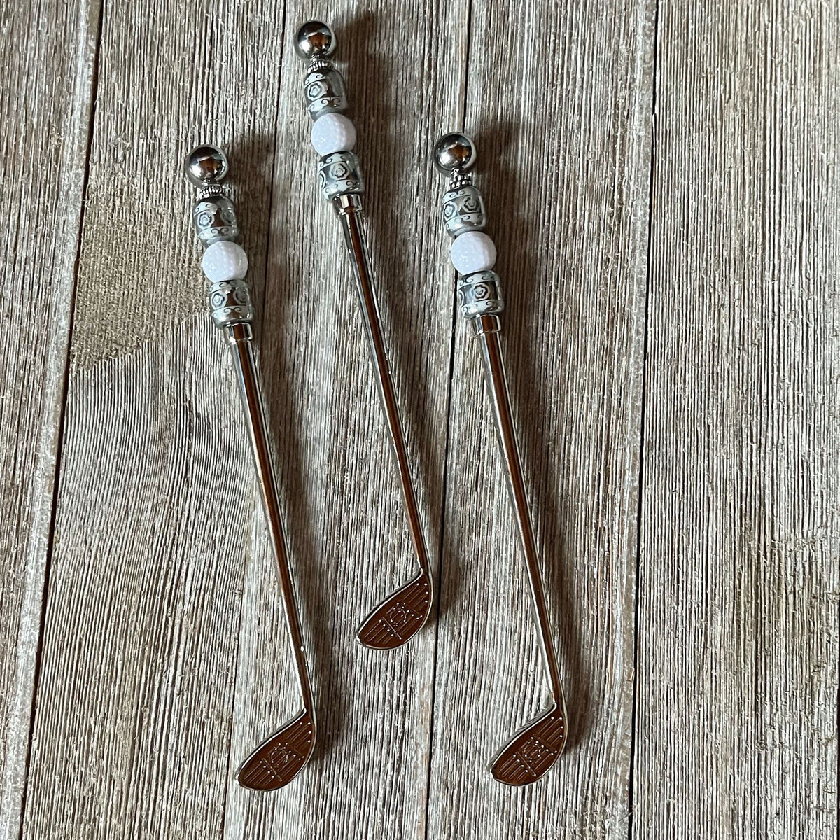 Set of three handcrafted golf ball stir sticks and spoon shaped like golf clubs, adorned with decorative beads, displayed on wood—a unique golfer barware gift for artisanal collections.