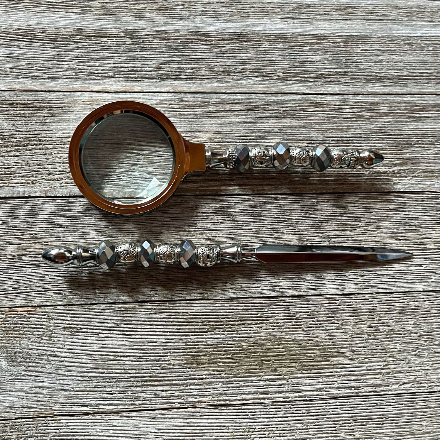 The Handmade Silver Desk Set features a magnifying glass and letter opener with faceted bead handles, shown here lying on a wooden surface.