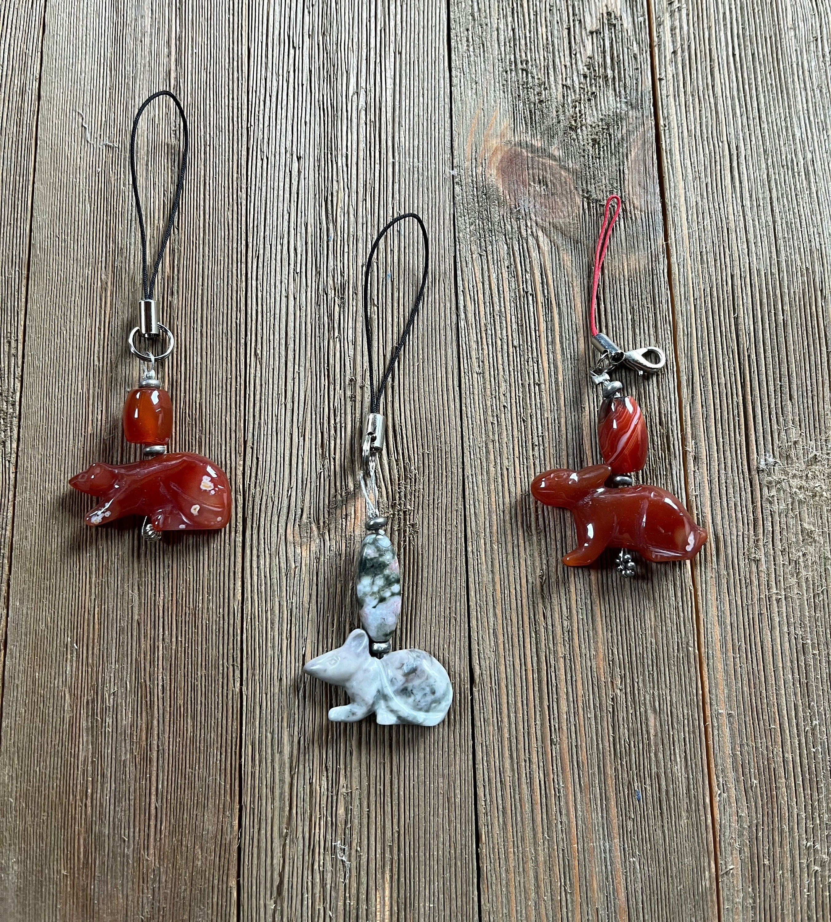 Two reddish-brown Carved Stone Animal Charms—one a Carnelian Rat, the other a Jasper Bunny Dangle—sit on string loops beside a grayish-white stone charm with dark patterns on a weathered wooden surface.