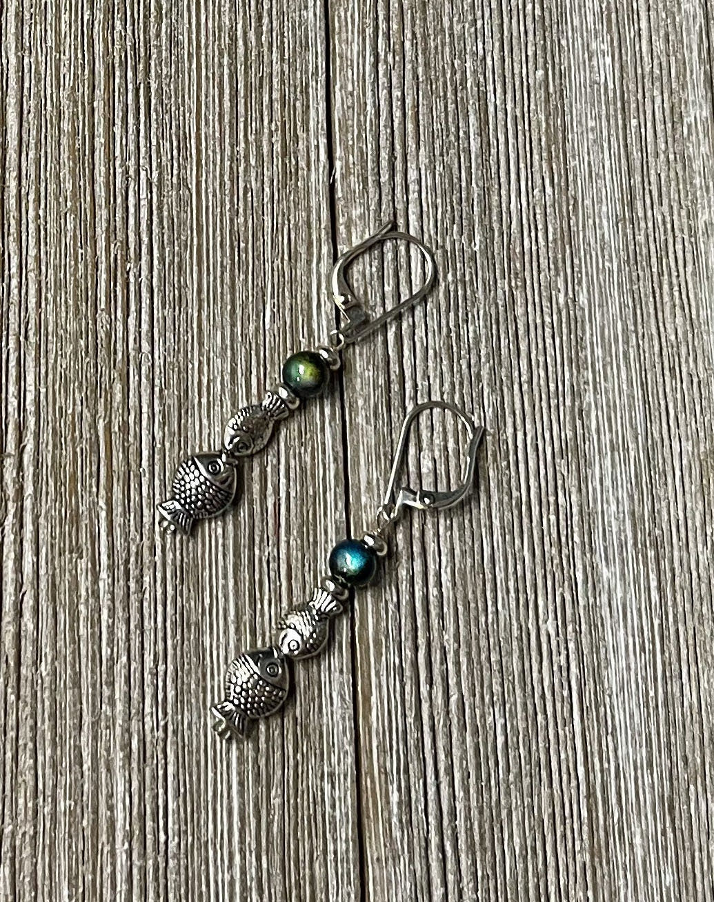 Fish Dangle Earrings by Gwynstone Handmade feature silver-tone fish charms and color-changing mood beads—perfect as an ocean lover gift. Displayed on a textured wooden surface.