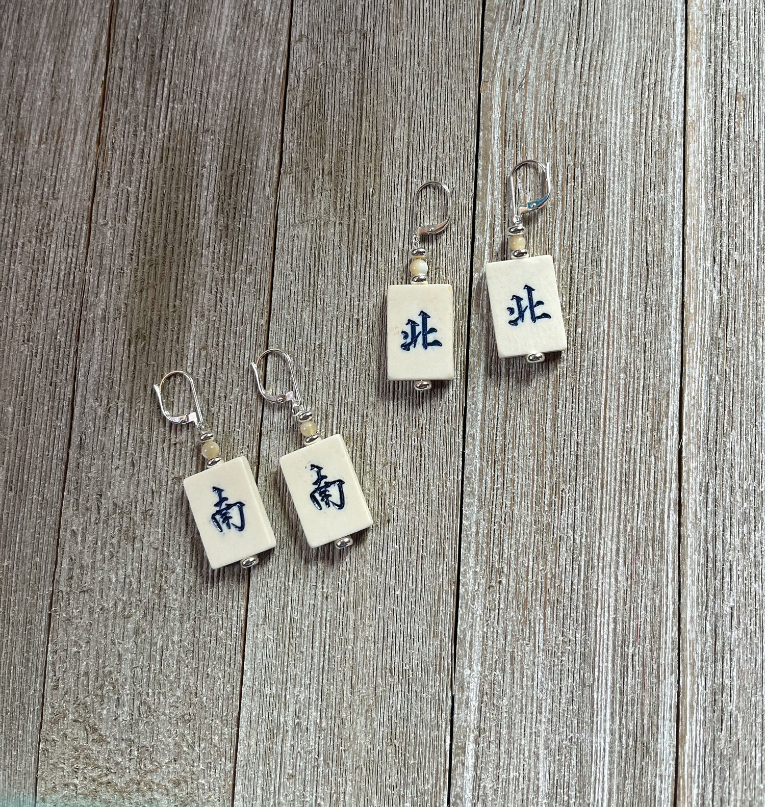 The Mahjong Tile Earrings: North/South Winds, perfect for mahjong players, feature four rectangular tiles with metallic hooks and black Chinese characters, arranged on a weathered wooden surface for a unique, Asian-inspired look.