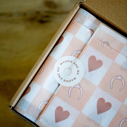 May include: A brown cardboard box with tissue paper featuring a pink and white checkered pattern with heart and headband illustrations. A round sticker with the text &amp;quot;MADDIE AND ME&amp;quot; and a headband illustration is on top.