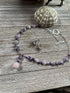 The Artisan Designed Jewelry Set features an amethyst and rose quartz necklace with sterling silver earrings, USA handcrafted, displayed on a rustic wooden surface next to a green sea urchin shell.