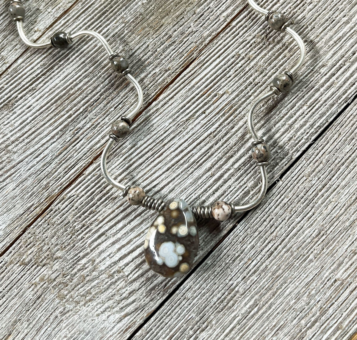 The Ocean Jasper Necklace Set: Artisan Wave Pendant Jewelry for Everyday Serenity by Gwynstone features handcrafted sterling silver with beads and a floral brown glass pendant, displayed on rustic wood.