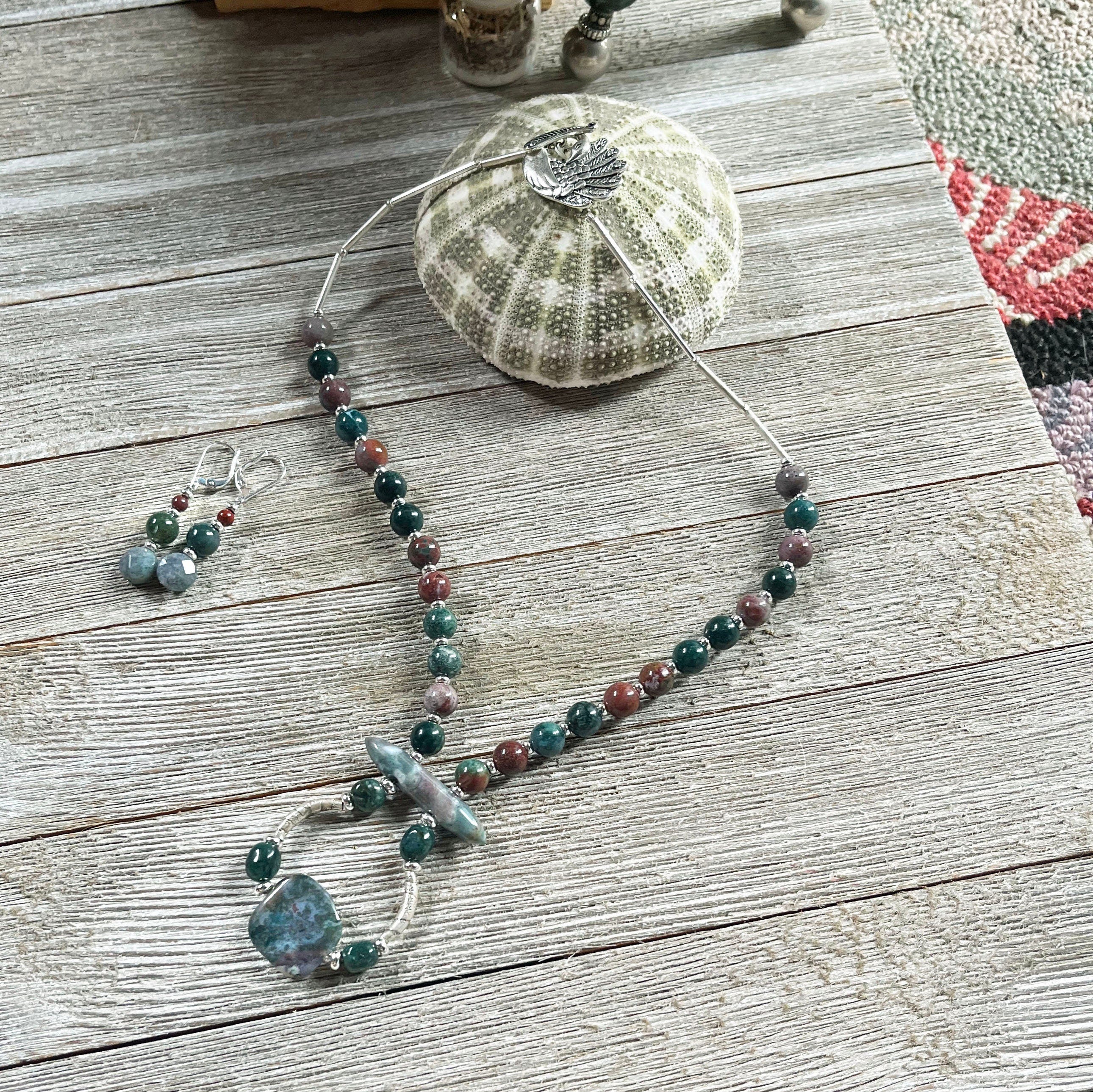 The Artisan Designed Fancy Jasper Jewelry Set with a sterling silver peacock clasp features a beaded necklace and matching earrings in green, brown, and blue tones, beautifully displayed on wood with sea-inspired accents.