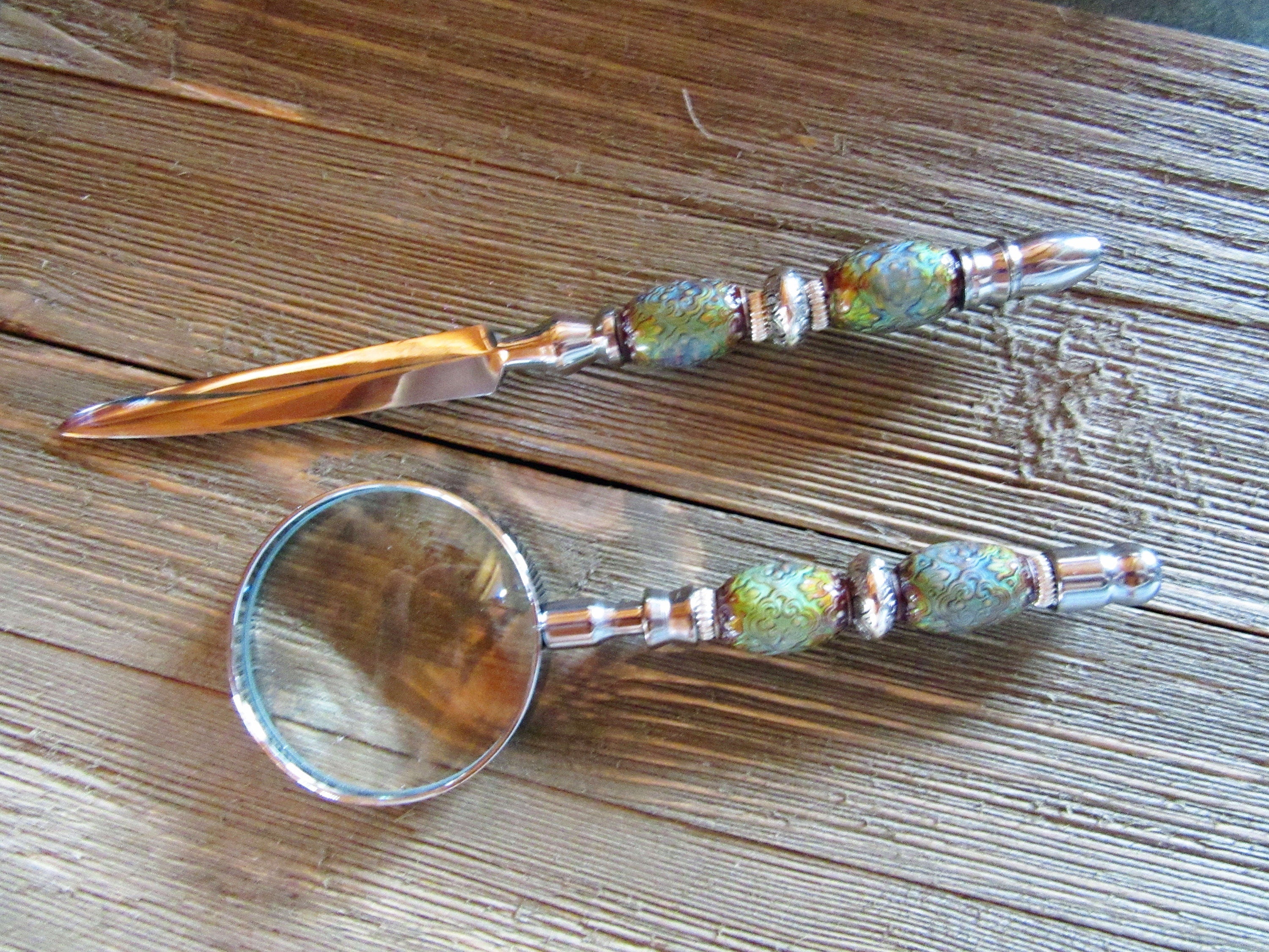 A Desk Set featuring a magnifying glass, letter opener, and pen with color changing beads rests on a wooden surface, combining elegance with a whimsical touch.