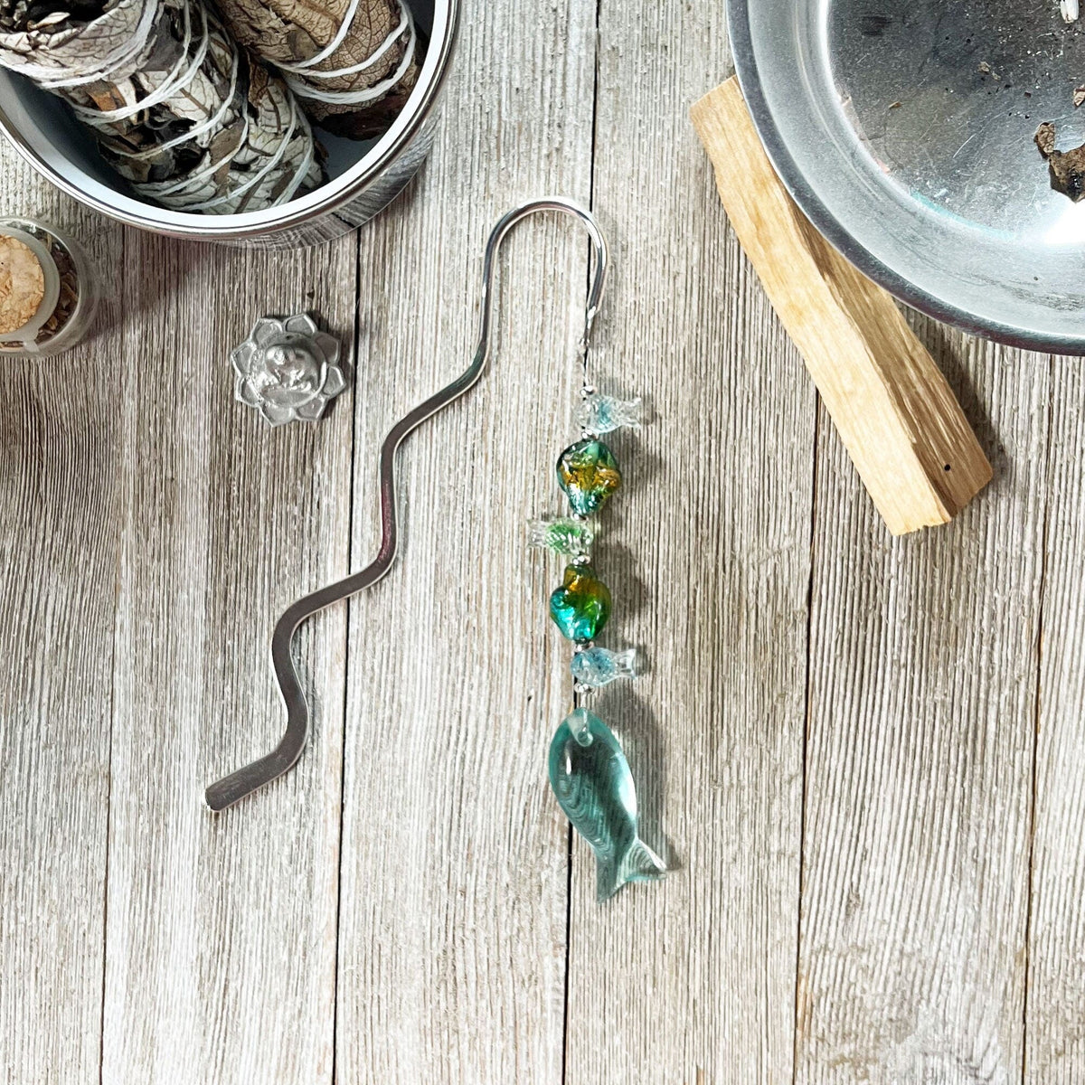 The Fish Stringer Bookmark—silver metal with green, gold, and clear beads finished by a glass fish charm—rests on light wood near sage bundles and palo santo. The perfect fisherman gift for spring!.