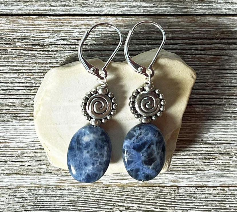 Artisan-designed Sodalite earrings with silver spiral accents, featuring dark blue oval stones, displayed on a pale seashell over rustic wood—unique gemstone jewelry for creativity, intuition, and self-expression.