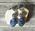 Artisan-designed Sodalite earrings with silver spiral accents, featuring dark blue oval stones, displayed on a pale seashell over rustic wood—unique gemstone jewelry for creativity, intuition, and self-expression.