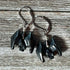 The Spooky Hematite Earrings for Halloween feature small black bat-shaped charms with spread wings and silver hooks, displayed on rustic wood. Perfect as good luck symbol creepy bat jewelry for the Halloween season.