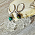 The Clear Dragon Earrings are handmade fantasy jewelry featuring color-changing quartz, iridescent dark green beads, and sterling silver hooks, displayed on rustic wood with a light rock in the background.