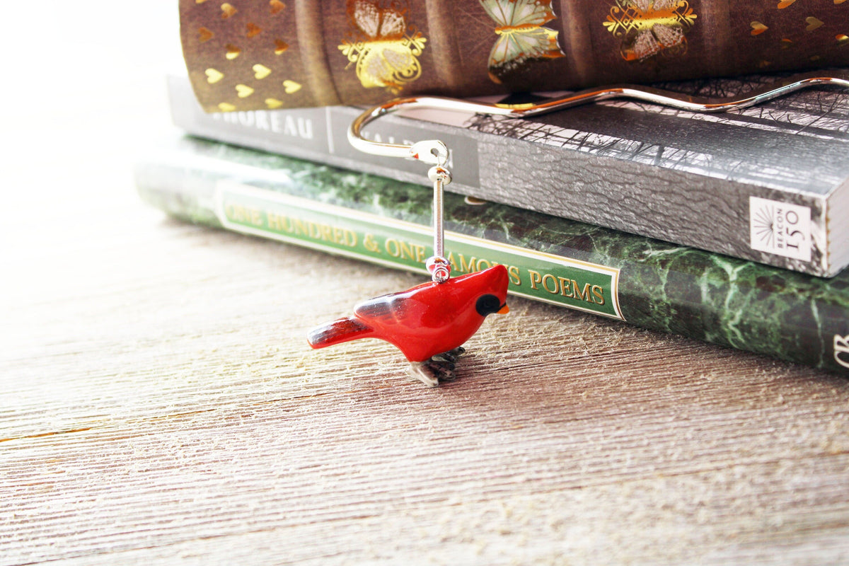 The Cardinal Bookmark features a porcelain red bird figurine on a silver metal bookmark, shown atop a green-covered poetry book and others on a wooden surface.