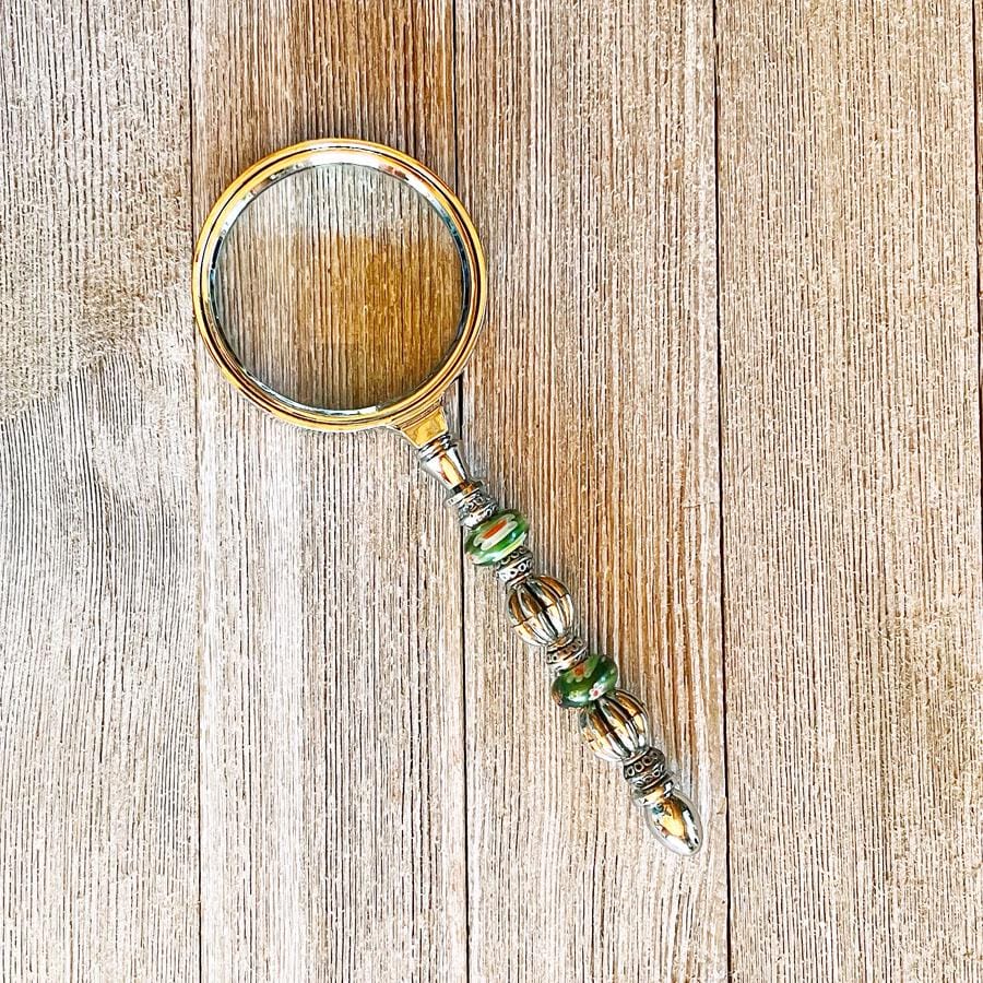 The Daisy Green Beaded Magnifier, a handmade silver office crafting tool with green accents, rests on a wooden surface displaying its unique beaded handle and elegant design.