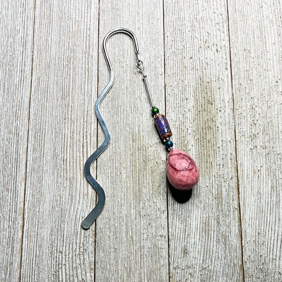 The Handcrafted Stone Bookmark: Teardrop Gemstone, with a wavy silver design and a pink stone charm accented by colorful beads, rests on light wood—a thoughtful gift for book lovers.