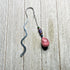The Handcrafted Stone Bookmark: Teardrop Gemstone, with a wavy silver design and a pink stone charm accented by colorful beads, rests on light wood—a thoughtful gift for book lovers.
