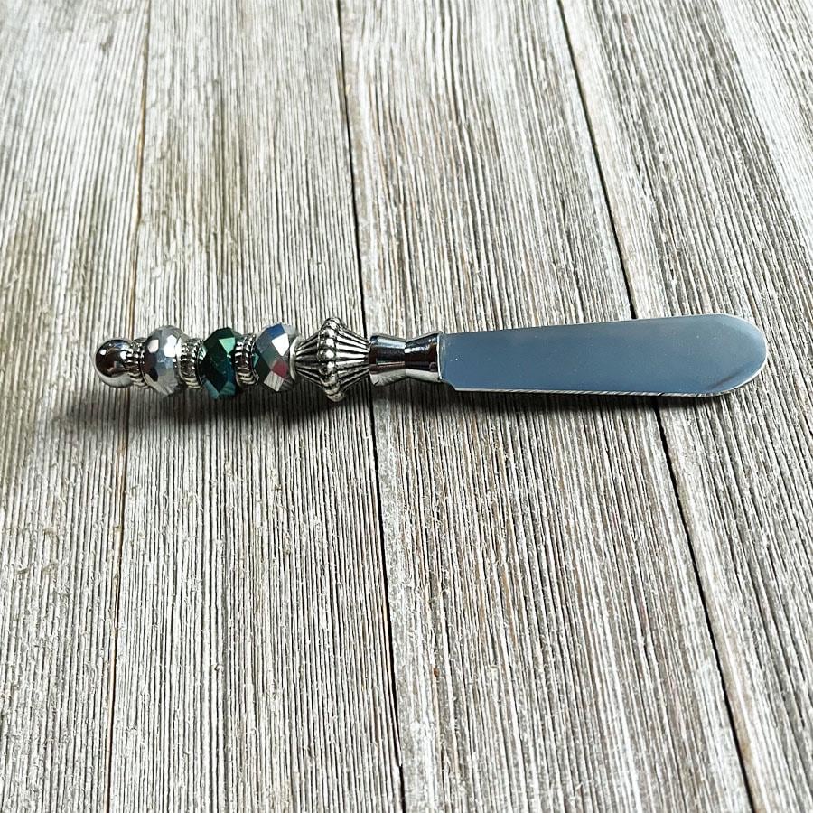 The Dark Faceted Glass Beaded Cheese Knife: Handcrafted Dip Spreader features a faceted glass bead on its metal handle, bringing elegance and style to any table setting when placed on a rustic wooden surface.