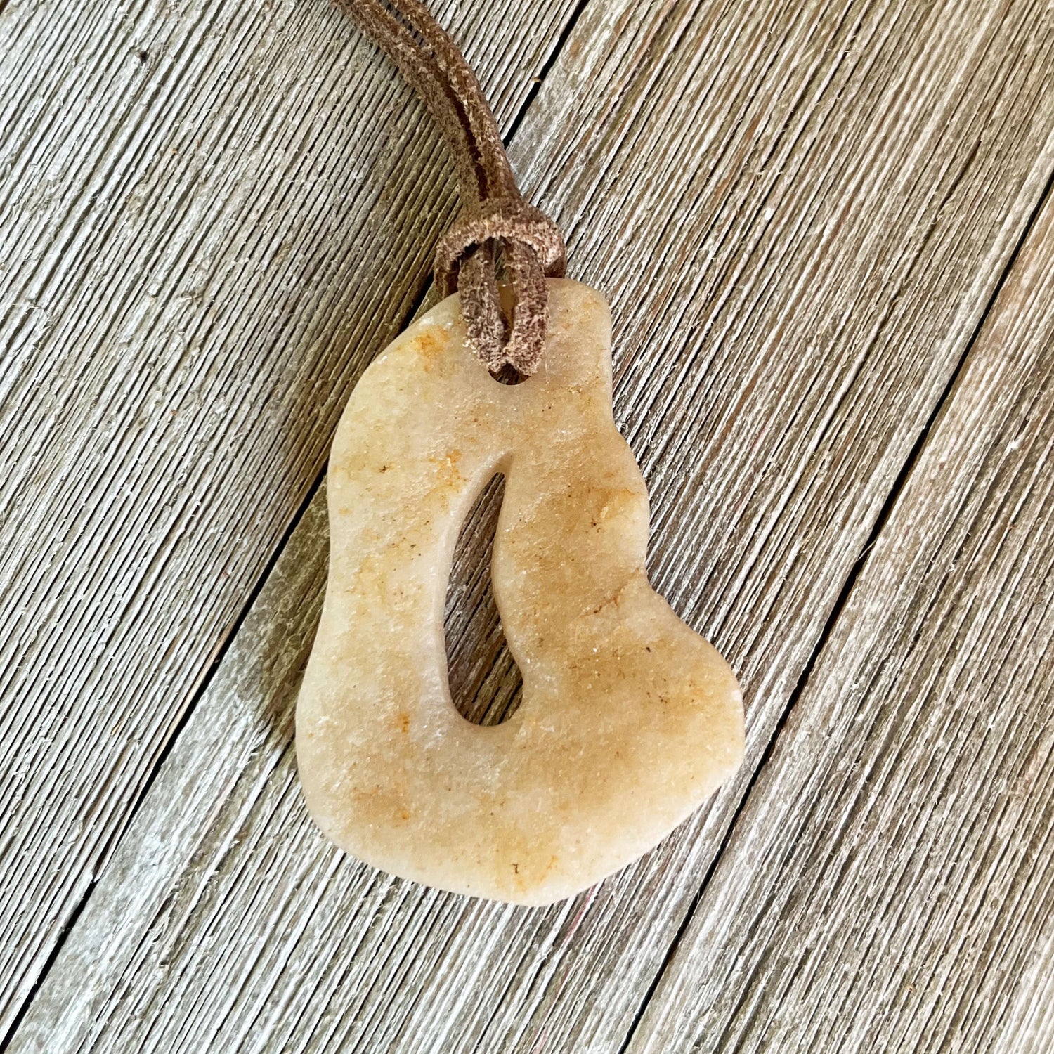 The Stone Pendant Necklace features a beige, freeform jasper pendant with a central cut-out on an adjustable leather cord, shown resting on rustic wood with visible grain.