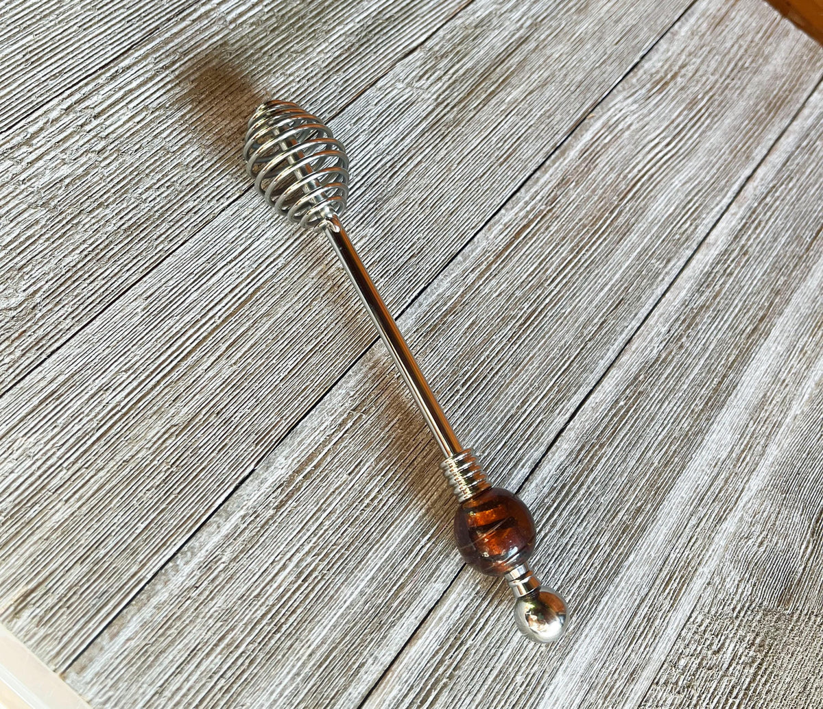 The Artisan Honey Drizzler features a spiral metal end and a brown &amp; golden art glass bead, measuring 6 inches, shown resting on a textured wooden surface.