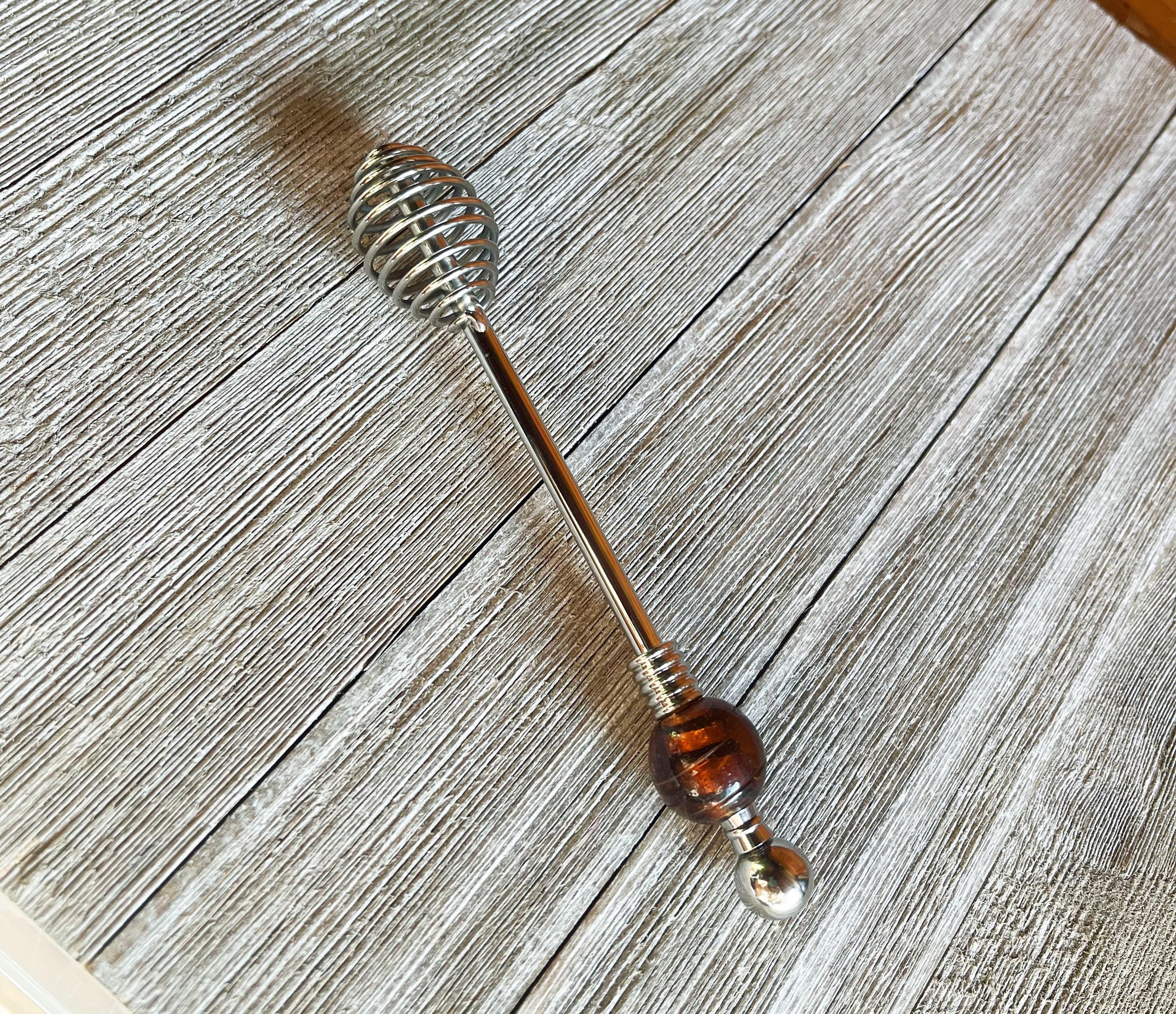The Artisan Honey Drizzler features a spiral metal end and a brown &amp; golden art glass bead, measuring 6 inches, shown resting on a textured wooden surface.