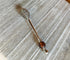 The Artisan Honey Drizzler features a spiral metal end and a brown & golden art glass bead, measuring 6 inches, shown resting on a textured wooden surface.
