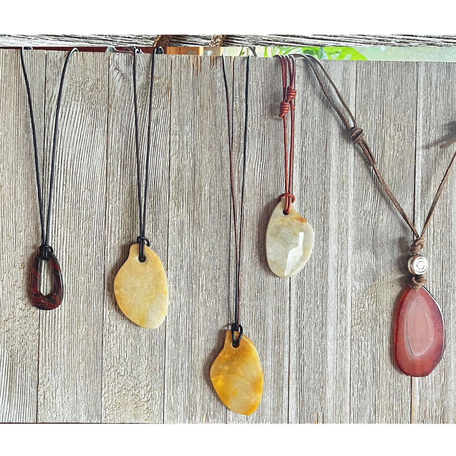 The Bold Leather Necklace with Stone Pendant features an adjustable cord and a unique earthy stone in varied shapes and colors—brown, yellow, cream, or red—displayed against a rustic wooden background.