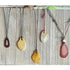 The Bold Leather Necklace with Stone Pendant features an adjustable cord and a unique earthy stone in varied shapes and colors—brown, yellow, cream, or red—displayed against a rustic wooden background.