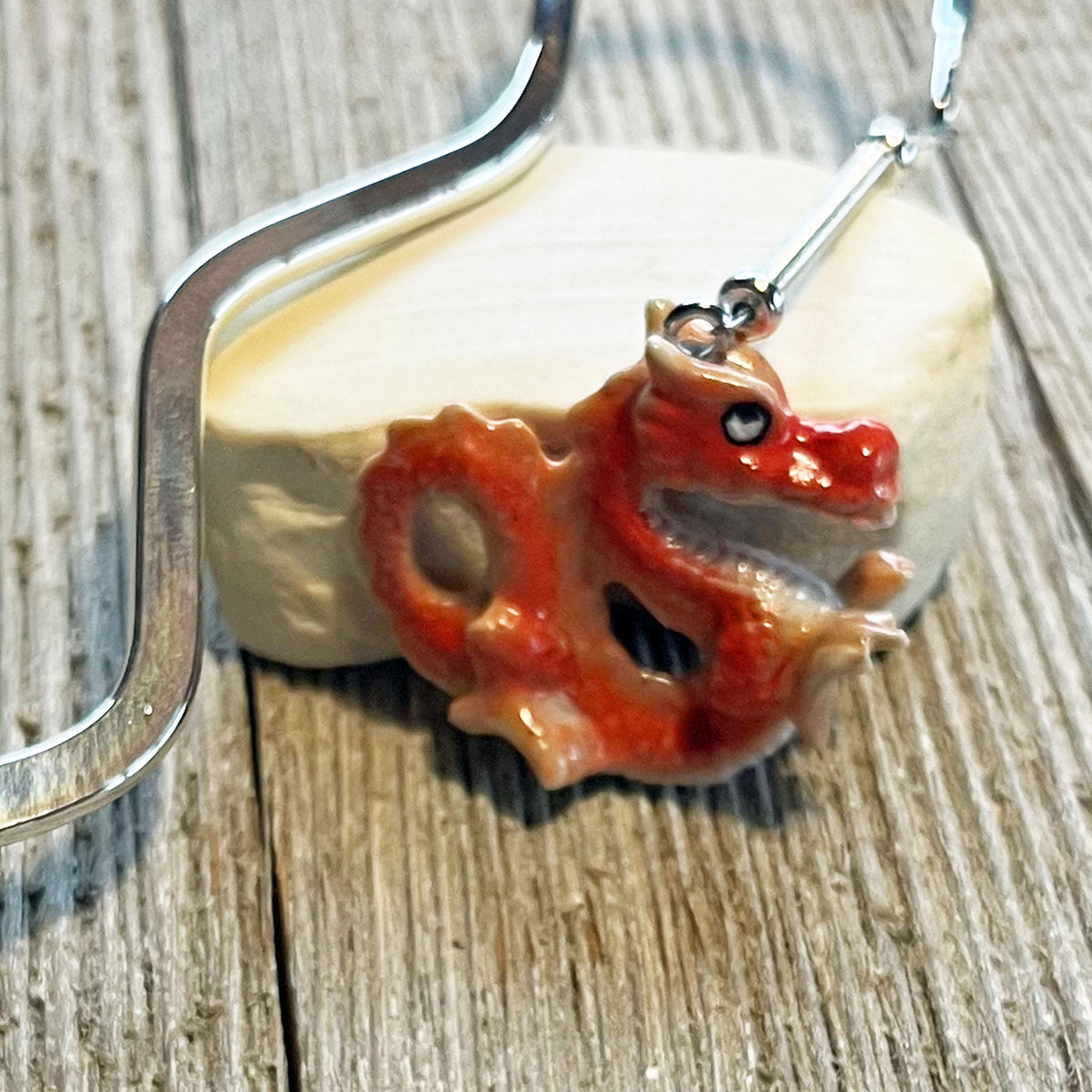 A close-up of the Red Dragon Bookmark—a handmade porcelain fantasy gift—shows its dragon-shaped pendant with a silver hook on a white oval stone, all set atop a textured wooden surface.