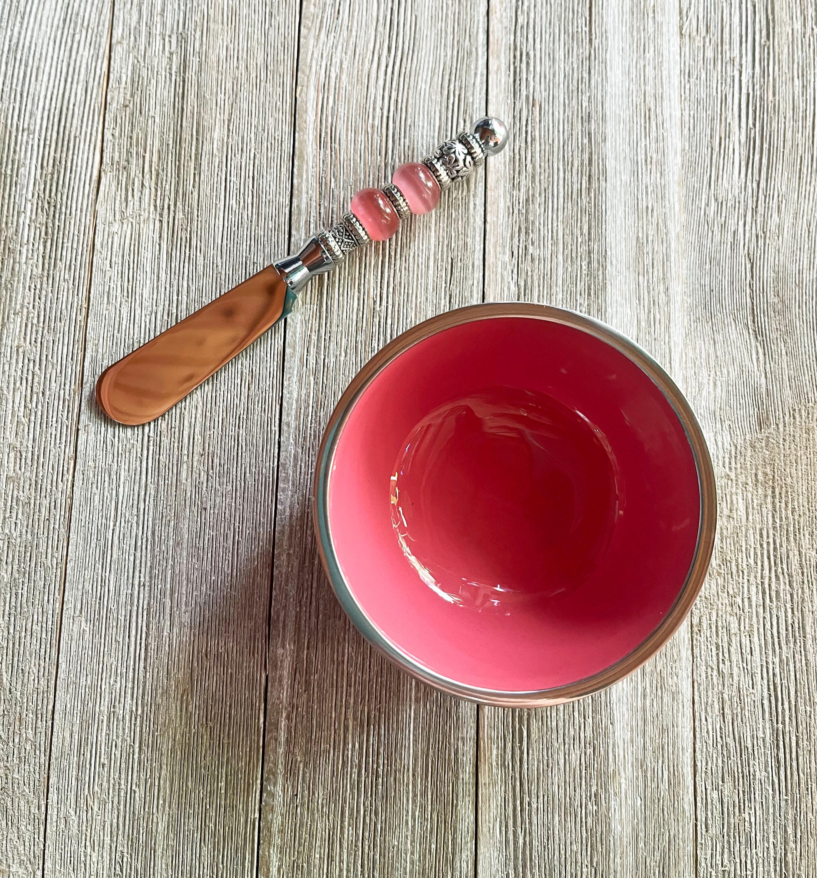The Pink Beaded Dip Spreader with Stainless Steel Bowl Set includes a small pink bowl with a silver rim and a decorative spoon featuring a beaded handle, displayed on a wooden surface.