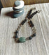 The Breciated Jasper Green Stone Necklace—an artisan-made essential oil diffuser with therapeutic lava beads and a minimalist design—rests on a wooden surface beside a small stack of balanced stones, making it a unique gift for him.